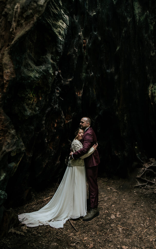 During their forest wedding, a bride and groom embrace in a hollowed out Redwood. The texture of the bark creates a beautiful visual as the soft light cascades down them