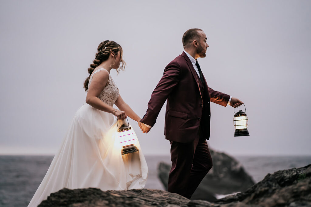 a bride and groom explore the rocky shore with lanterns in hand