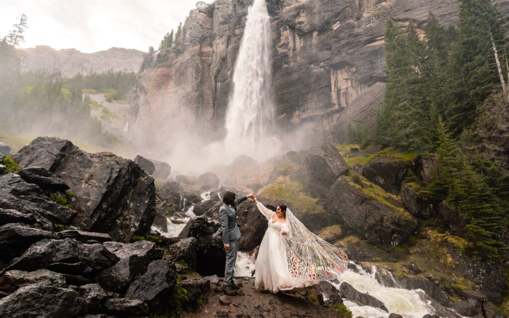 Couple in wedding attire dances in front of a gushing waterfall during their sunrise elopement