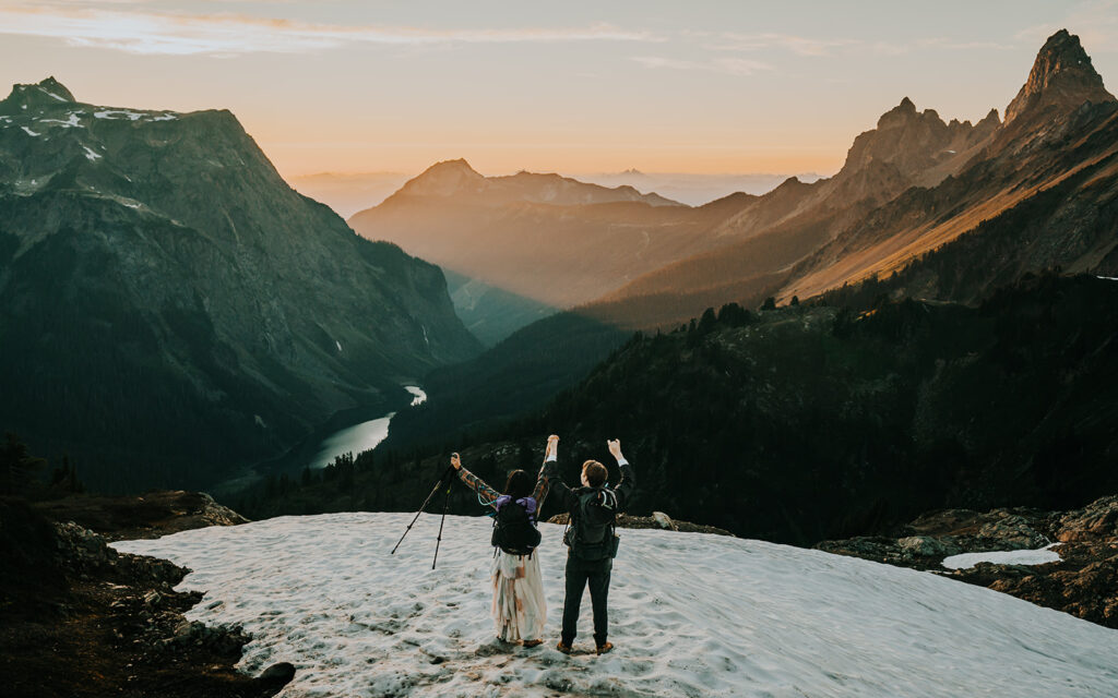 a bride and groom in their wedding attire stand on a snowfield and cheer into the unending mountain range painted in sunset tones during their hiking elopement
