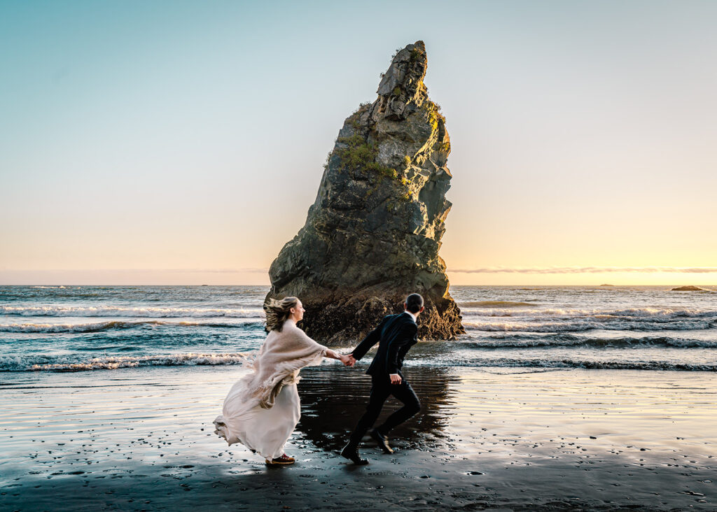 a couple in their wedding attire run in front of a gorgeous sea stack illuminated by the sunset. the waves are a pinky blue.