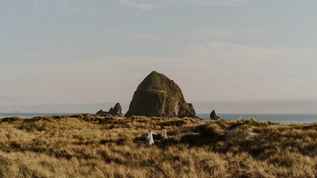 a sweeping landscape shot of the iconic haystack rock on cannon beach. Seagrass sways in the foreground as a bride leads her partner from the beach. 
