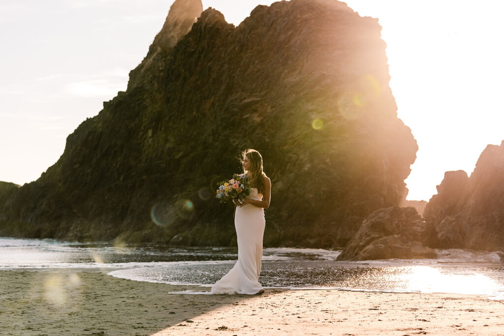 a wide shot of a bride standing in front of a large sea stack. the golden light of the sunset creates a glowy light and lens flare 