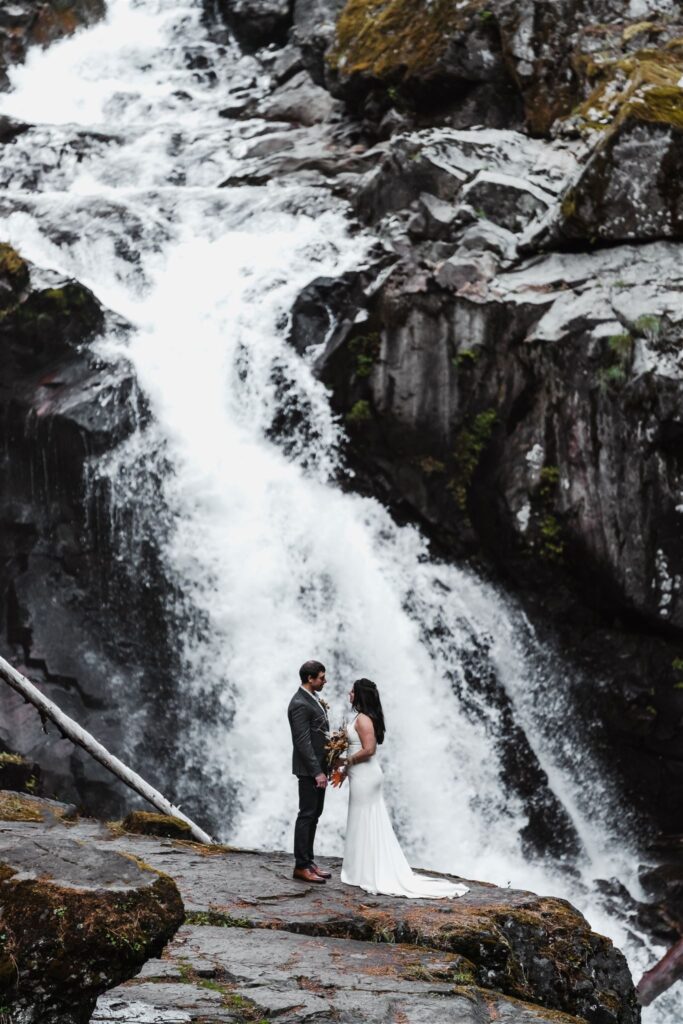 after they decided how to elope, a bride and groom stand in front of a cascading waterfall in their wedding attire 