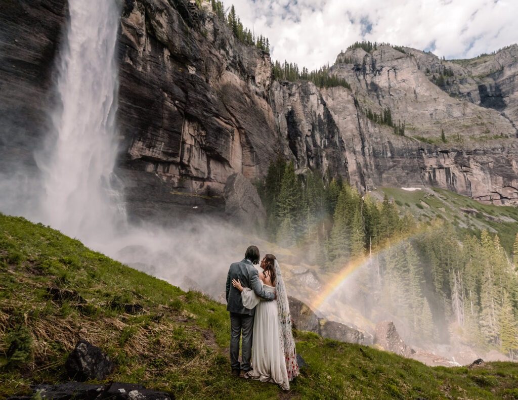 after they decided how to elope, a bride and groom admire a gushing waterfall and stunning rainbow