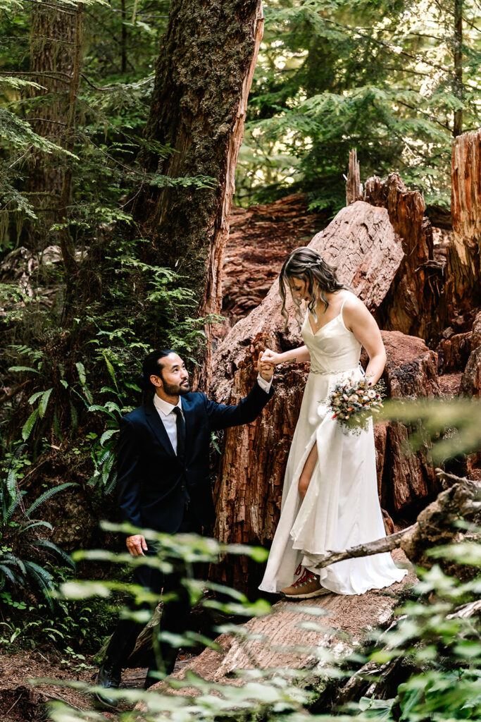 after they decided how to elope, a groom helps his bride hop down from a fallen tree in a lush, green forest 