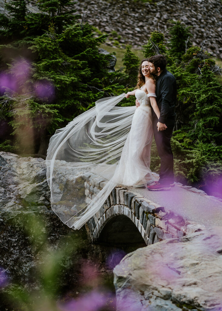 a couple embraces on a stone bridge. The groom embraces his bride as she swirls her sheer train in the wind. The are framed by purple wildflowers