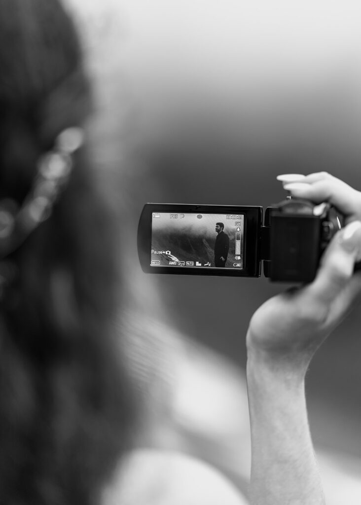 a black and white image taken over the shoulder of a bride as she films her groom on a handheld camcorder 
