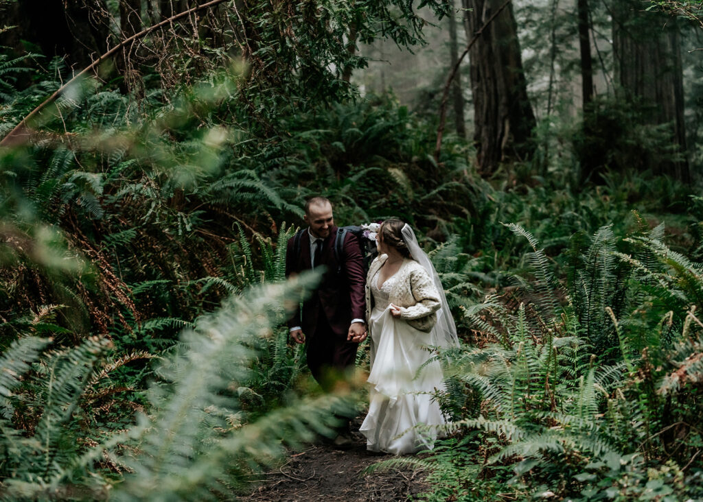 after they decided how to elope, a bride and groom walk through a lush green forest. They smile widely at each other as they explore