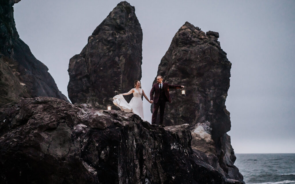 after they decided how to elope, a bride and groom stand on the edge of a sea cliff. they hold lanterns as they explore in twilight, in their wedding attire 