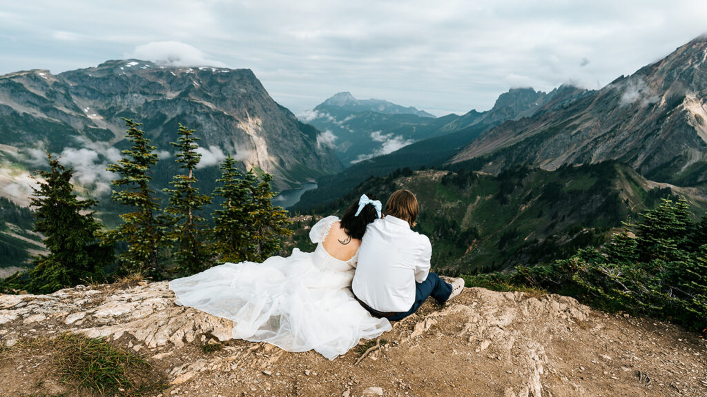 a couple sits on the edge of a cliff in their wedding attire. they snuggle as they gaze at the sweeping mountains that sprawl before them 