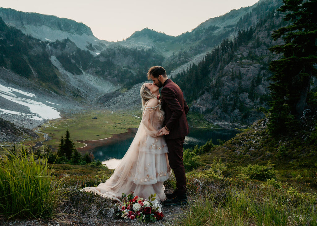an ethereal scene of a bride and groom kissing in a beautiful  blue and green landscape. A lake ca bee seen beneath them and the mountains rise above them.
