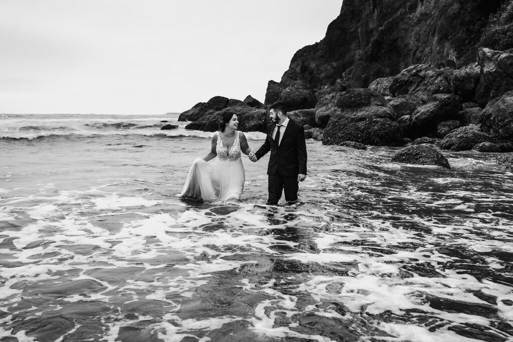 after they decided how to elope, a bride and groom smile at each other as they run through the icy waters of the pacific. Rocky sea stacks pop out behind them