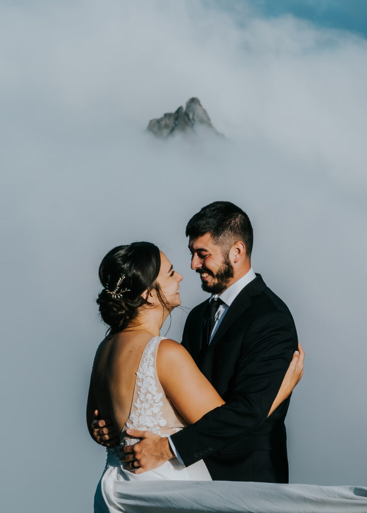 a bride and groom embrace and smile widely at each other. fog rolls through the landscape and just the very top of a mountains peeks out through the mist