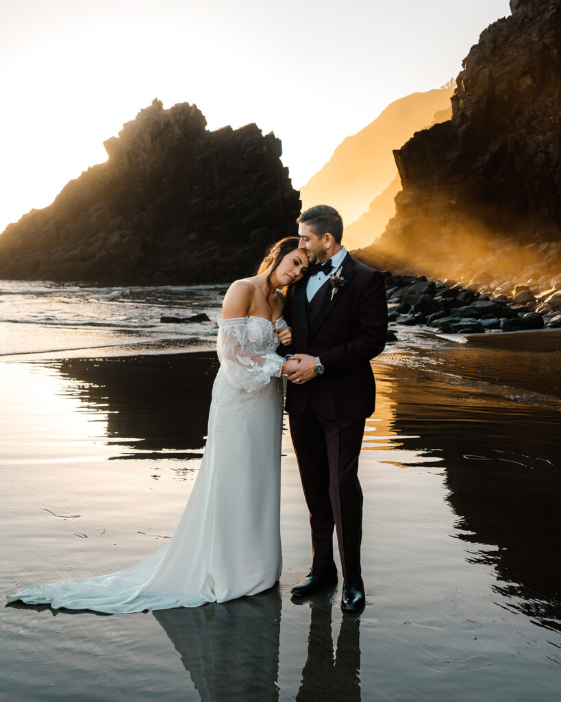 after they decided how to elope, a bride and groom stand on a rocky beach. she rests her head on his shoulder as golden light pours through the sea stacks 