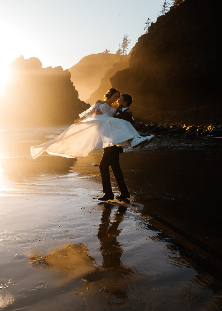 after they decided how to elope, a groom lifts his bride and spins her in the golden light of the sunset. her dress billows as he twirls here in front of basalt sea stacks 