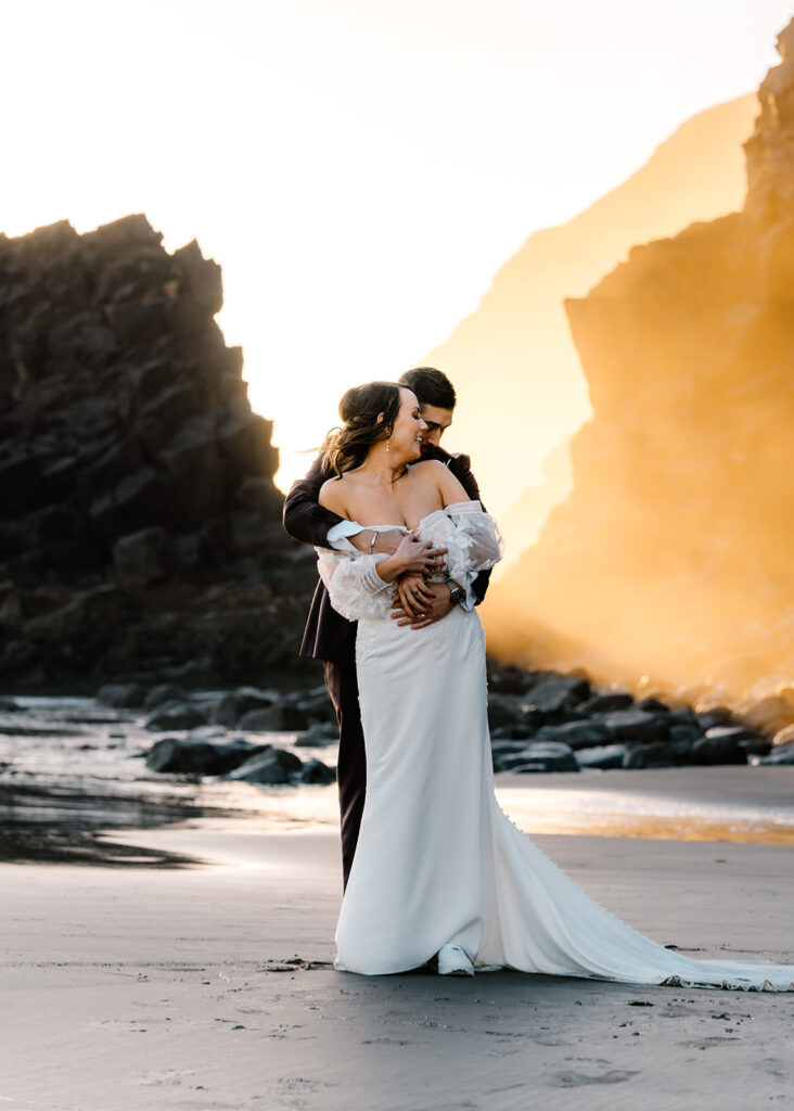after they decided how to elope, a bride and groom embrace on a rocky beach. glowy sunlight seeps through the space between the dark sea stacks
