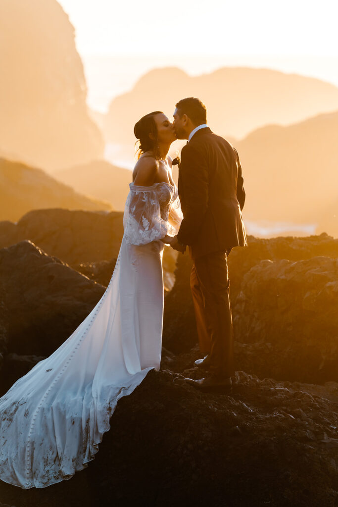 after they decided how to elope, a bride and groom stand on a rocky beach and kiss as the sun sets behind them 