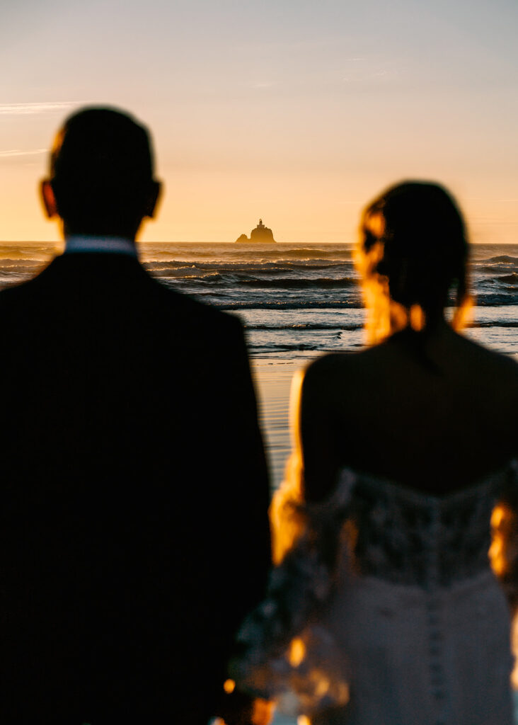 after they decided how to elope, they gaze out at a distant lighthouse. they frame the structure as they drink in the view in their wedding attire 