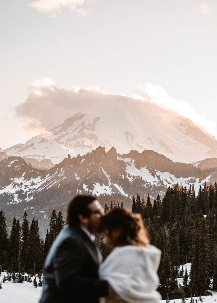 a clear shot of a glowy, mount rainier. a blurred out couple, in their wedding attire embrace in the foreground. the groom kisses the top of his brides head
