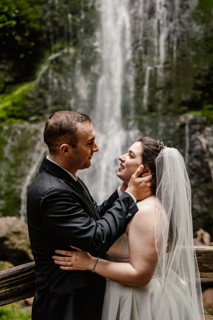a groom takes his brides face in his hands. They gaze lovingly at each other in front of marymere falls preparing for their lake crescent wedding.
