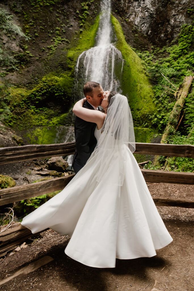 a bride and groom kiss in their wedding attire in front of marymere falls before their lake cresent wedding 
