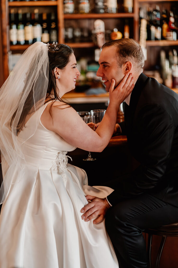 a bride and groom sit at a beautiful bar and share a glass of champagne together before their lake crescent wedding. She smiles broadly and touches her grooms cheek as he gazes at her.