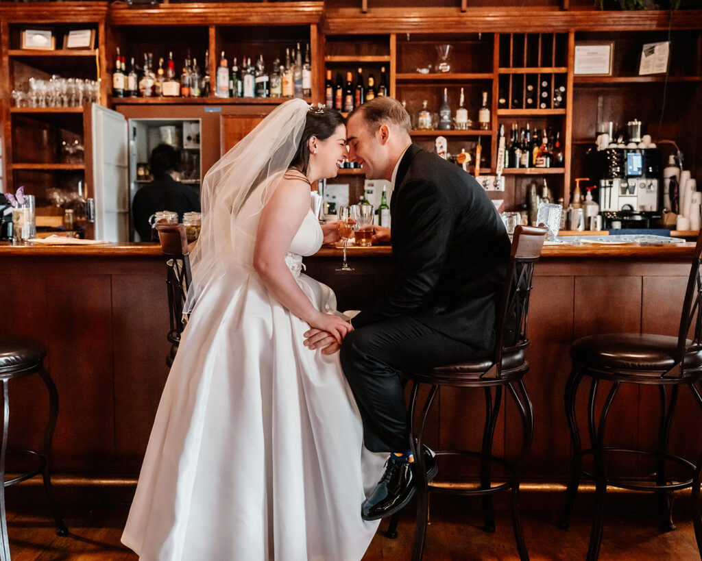 a bride and groom in their wedding attire sit at a wooden bar and share a drink to celebrate their lake crescent wedding