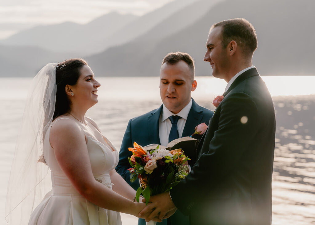 a bride and groom smile sweetly at each other during their lake crescent wedding ceremony. light bounces off of the lakes surfaces creating a dreamy ambience.