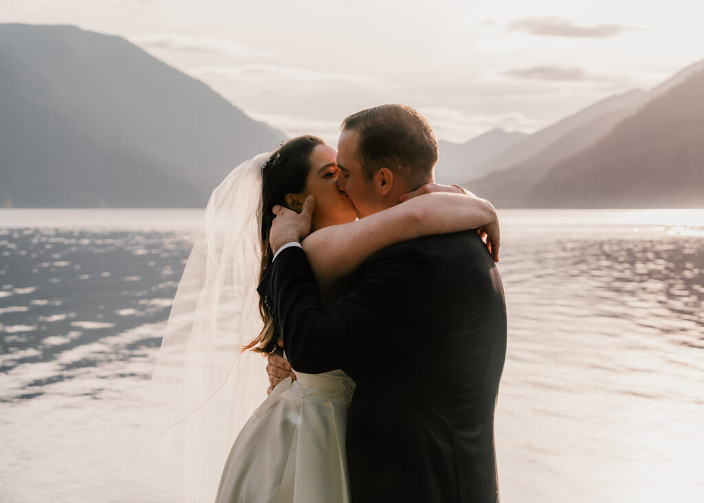 a bride and groom share their first kiss during their lake crescent wedding. The sun has begun to set and rose gold light reflects off of the blue waters of the lake 