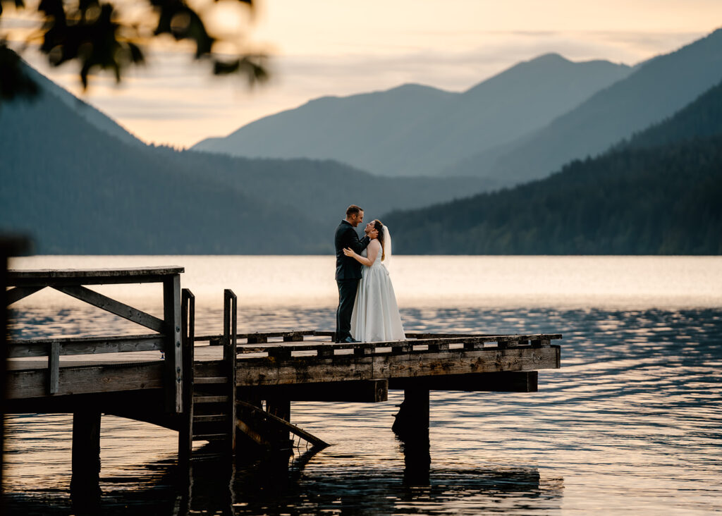 a couple in their wedding attire embrace at the edge of a wooden dock during the sunset of their lake crescent wedding. The blue mountains roll behind them, disappearing into the rose gold waters of the lake. 