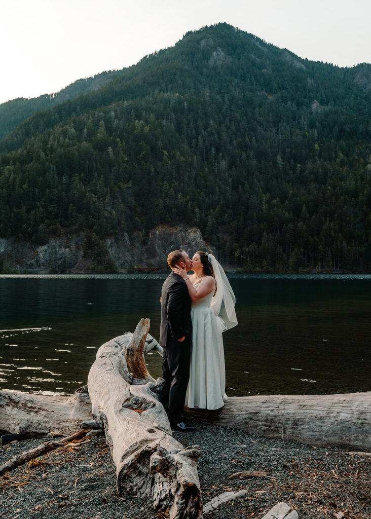 A bride and groom kiss after their lake crescent wedding. she stands on a large piece of driftwood, matching his height. With his face in her hands, she kisses him.