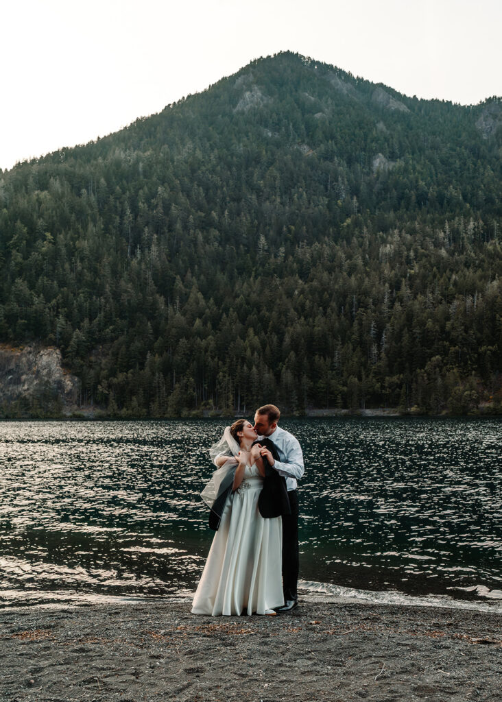 a bride and groom embrace in front of barnes point. The waters of lake crescent ripple behind them. the groom wraps his bride in his jacket to keep her warm. 