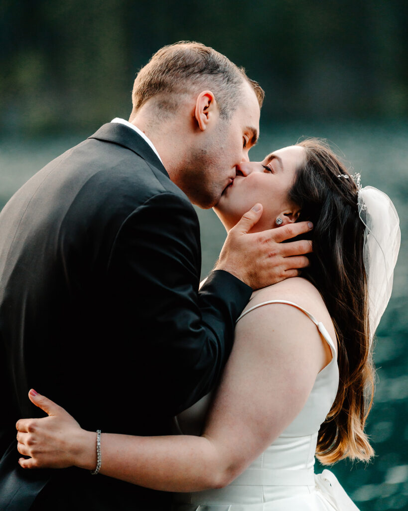 a bride and groom share a kiss. His hand rests on her cheek and the soft blue waters of lake crescent ripple behind them