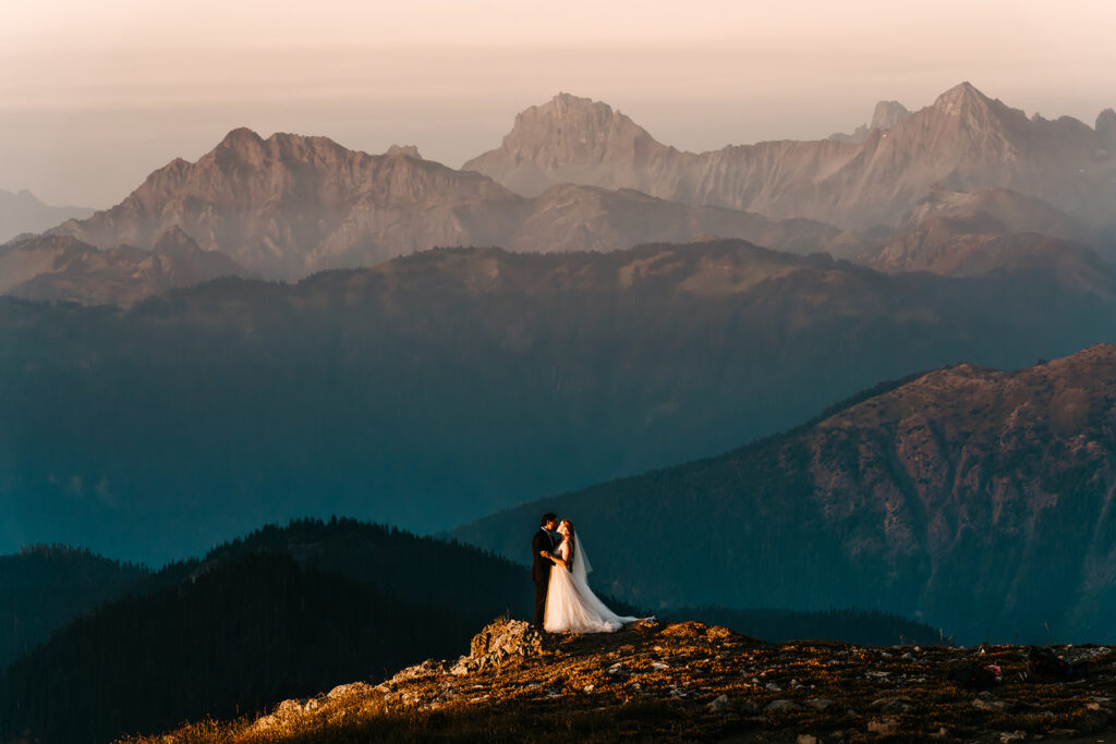 A couple embraces in their wedding attire as the sun sets on their last minute elopement. They are surrounded by layers of mountains that are layered with different hues of blue and pink