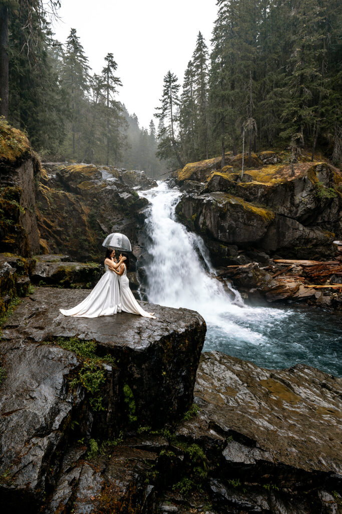 two brides stand cliffside in front of a gushing waterfall during their last minute elopement. It is raining lightly and they embrace under a clear umbrella