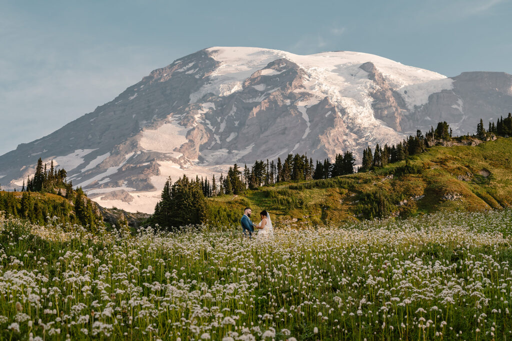 a bride and groom exchange vows in front of Rainier. The bottom third of the frame is filled with wildflowers, and the sun is shining golden hues on everything it touches during this last minute elopement