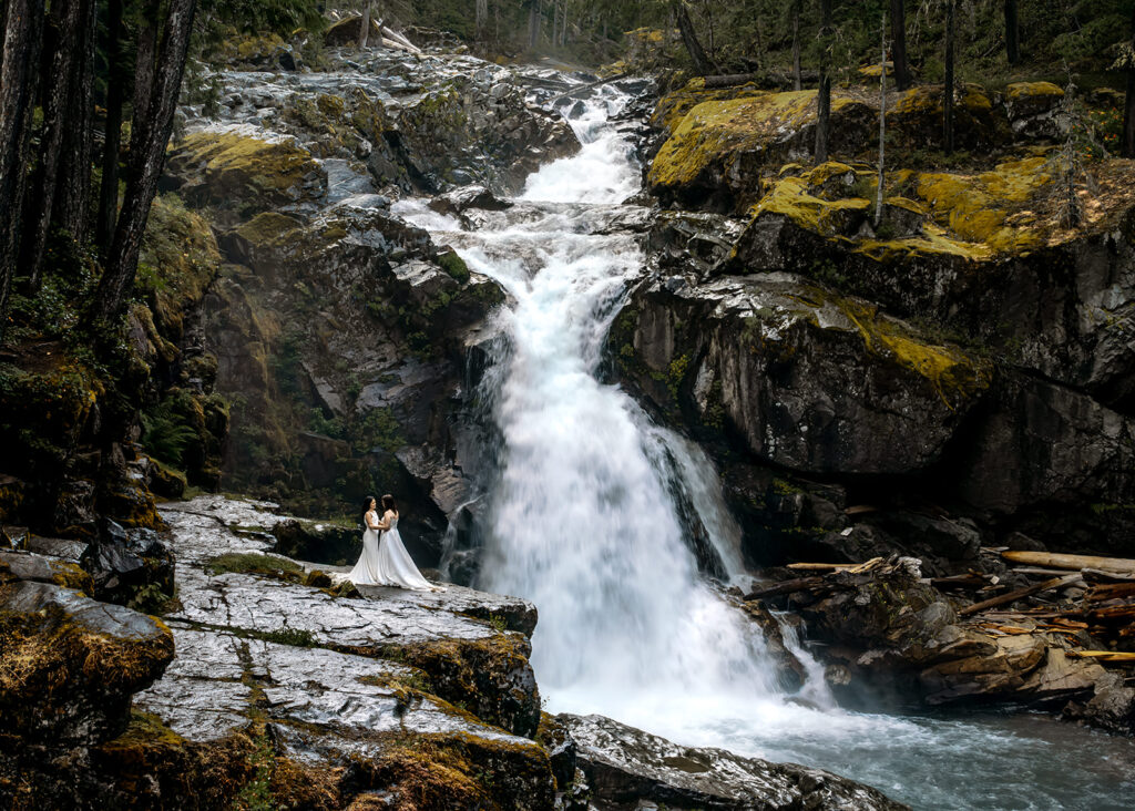 two brides embrace in front of a cascading waterfall during their last minute elopement. We see the falls flowing behind them as they embrace on a cliff. We see clear blue waters churning below them.