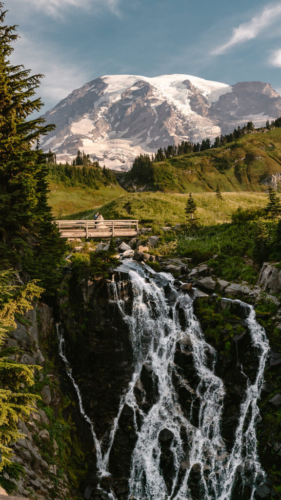 a couple in their wedding attire embrace over myrtle falls with Rainier in the background