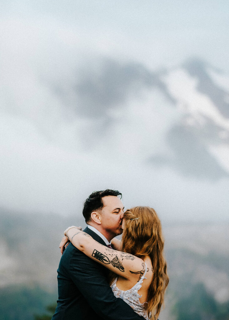 a groom kisses his bride on the forehead. the outline of a mountain can be seen behind them, barley peeking out behind the clouds.