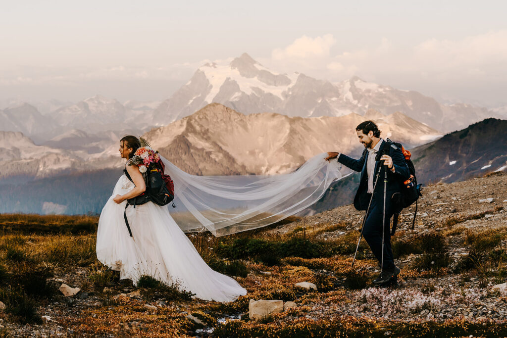 a bride and groom hike in their wedding attire during their last minute elopement. she leads the way, with her bouquet in her backpack. He carries her veil and trekking poles. Mountains sprout out behind them.