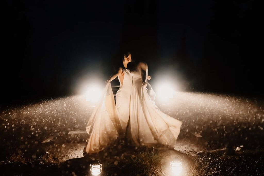 two brides dance in the rain, lit only by the headlights of a truck to celebrate their last minute elopement. Rain sparkles like glitter around them as they dance and kiss.