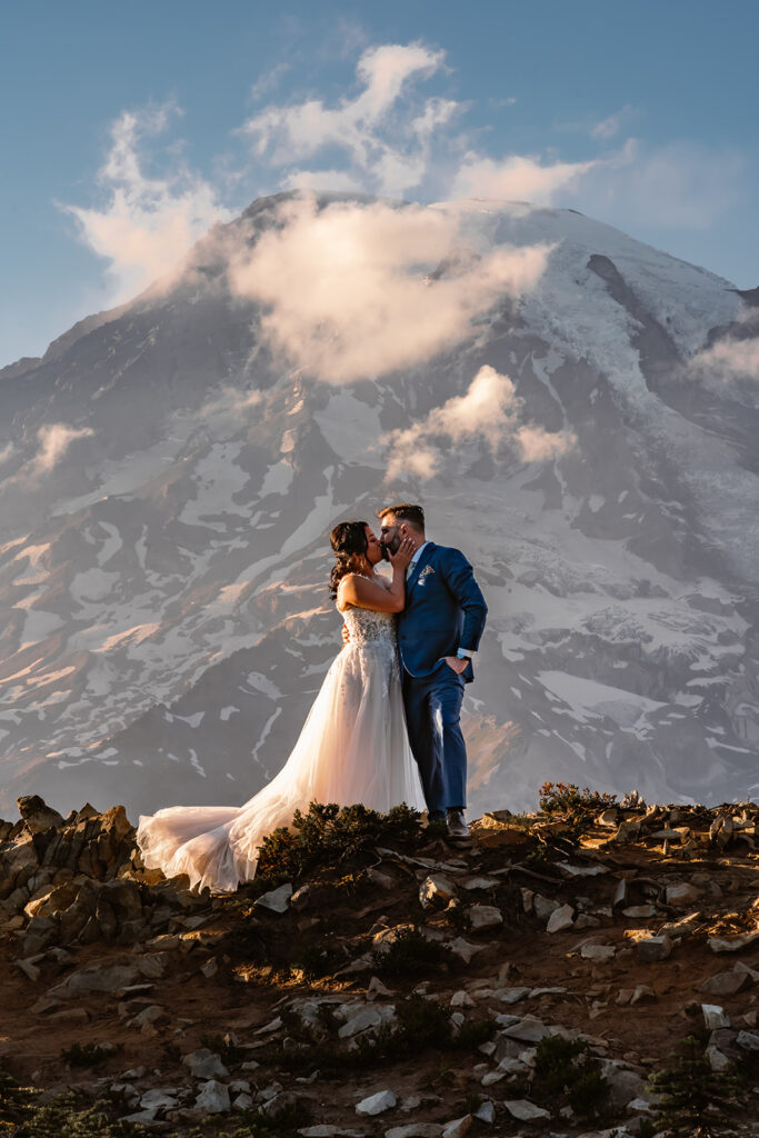 A bride and groom kiss in front of a towering mount rainier, one of the best places to elope in Washington. Golden, pink light highlights them and they are centered perfectly under a tiny puff of a cloud. 