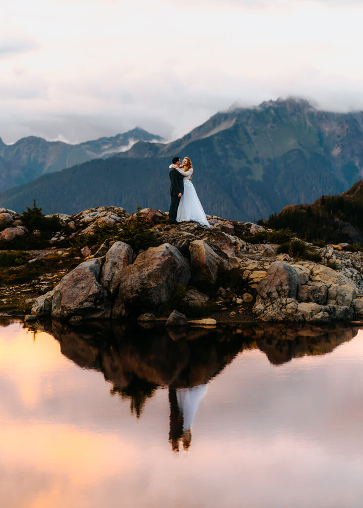 a bride and groom embrace on a large boulder. they are reflected in a mountain tarn below them, where we also see the pink and gold clouds from the sunset reflected back at us