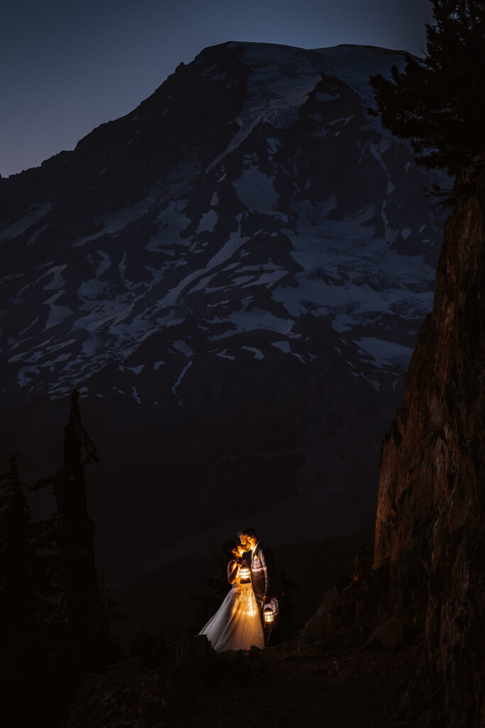 A couple in their wedding attire embrace in lantern light. a pale mount rainier stands behind them