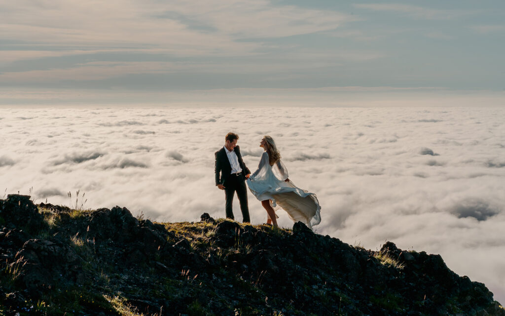 a brides dress billows in the wind as her groom admires her. they stand on the top of a mountain, a cloud inversion swirls below them 