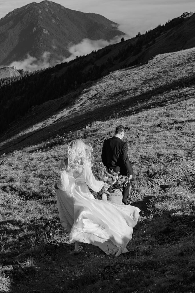 a bride follows her groom down a switchback, her wedding dress billowing in the wind during their Olympic Peninsula elopement. 