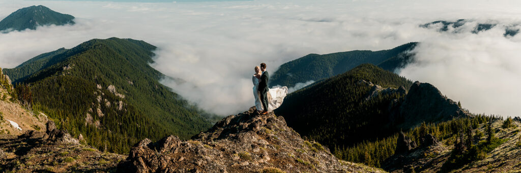 a panoramic image of a couple in wedding attire standing on top of a mountain during their oplympic peninsula elopement. mountains rise above them, a cloud inversion swirls below them. It is peak adventure romance.