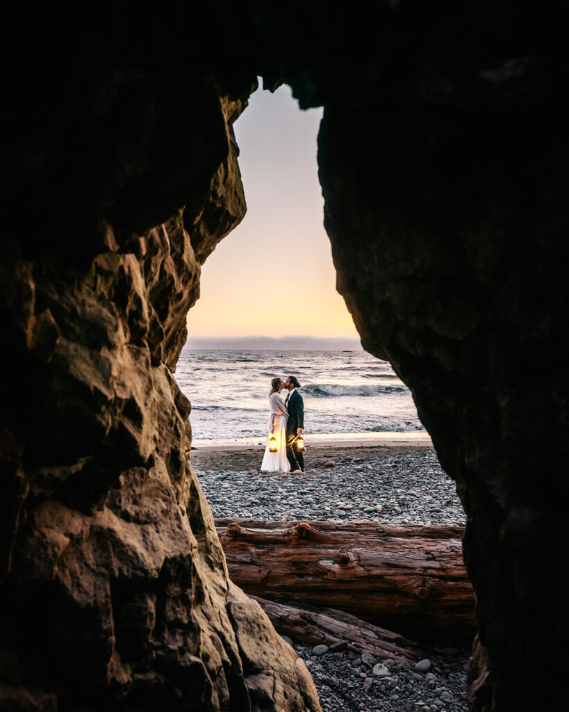 a couple in their wedding attire finish exploring one of the best places to elope in Washington by sharing a kiss by lantern light on a rocky beach. the scene is framed by a rocky seastack and illuminated by a punchy sunset 