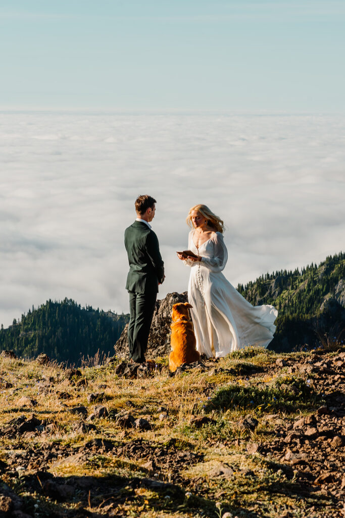 a bride reads her vows to her groom sanding on a mountain above a cloud inversion. their dog sits between them 
